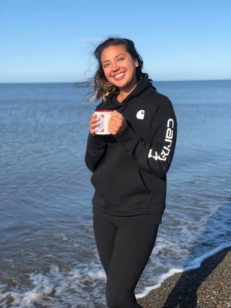 Full shot of Denali by the ocean holding a coffee mug